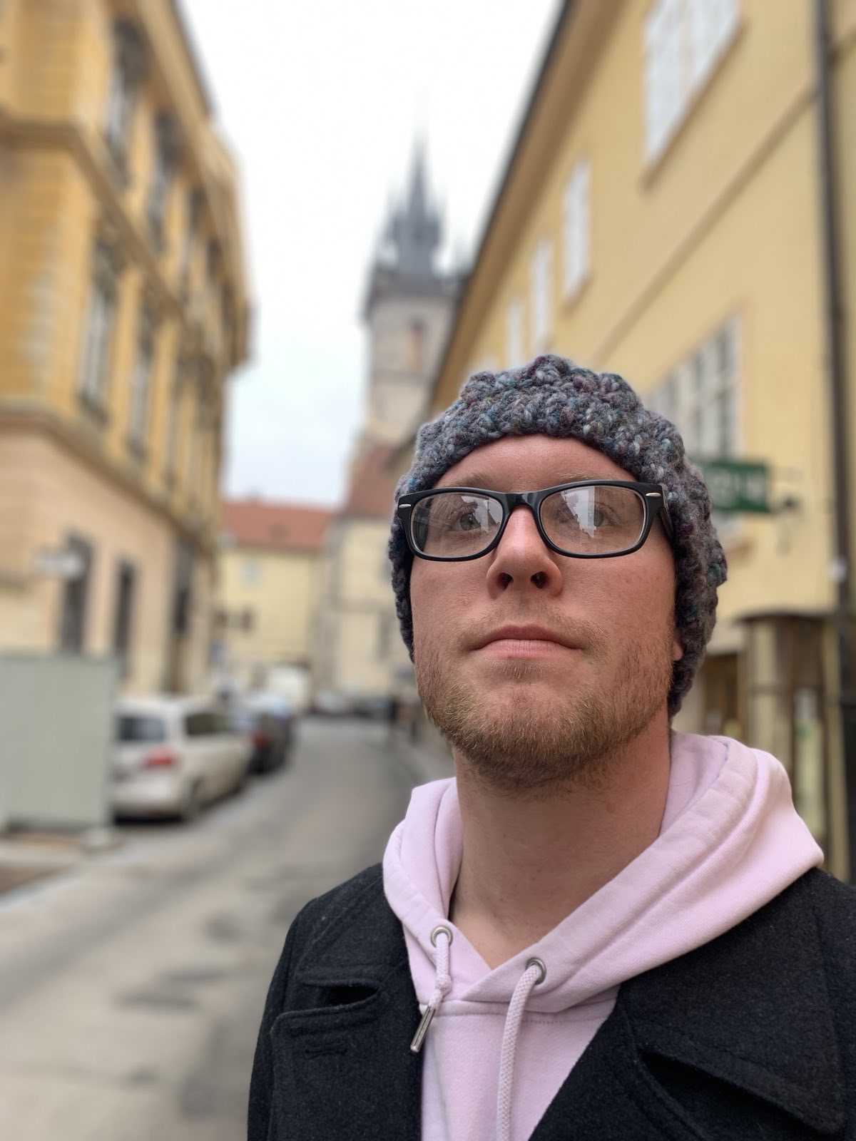 Image of a male with glasses in a sweater and beanie, looking beyond the camera with a cityscape out of focus behind him.